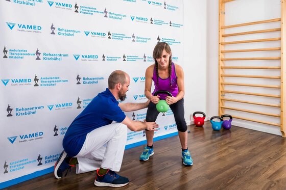 Eine Frau trainiert mit einer Kettlebell unter Anleitung eines Therapeuten in einem hellen Raum.