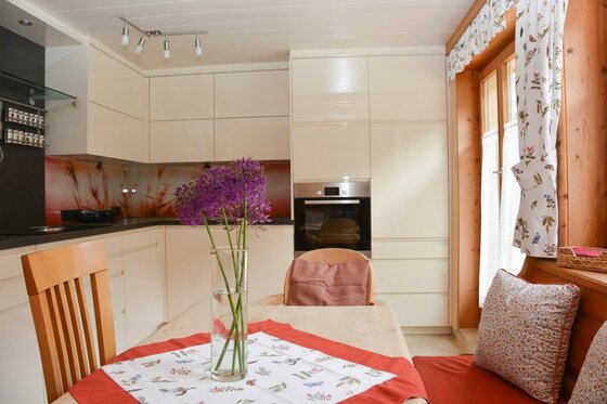 Moderne Einbauküche in Beige mit Holztisch, lila Blumen in einer Vase und einer Eckbank mit Kissen.