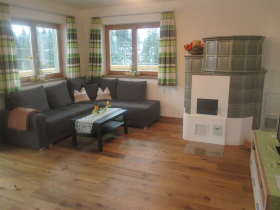 Wohnzimmer mit dunklem Ecksofa, Couchtisch, Kachelofen und Holzboden vor zwei Fenstern.