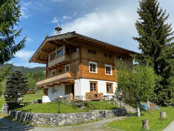 Haus Lazar im Sommer Dreistöckiges alpines Holzhaus mit weißem Erdgeschoss, Balkonen und Steinfundament vor Nadelbäumen.