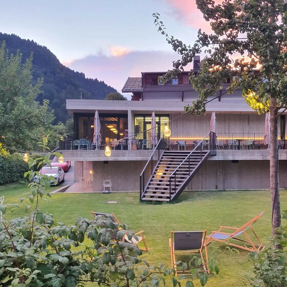 Gasthof Auwirt Außenansicht Modernes Gebäude mit Außenterrasse, Treppe und Liegestühlen auf einer Wiese vor bewaldeten Bergen.