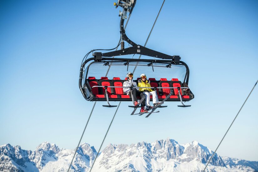 Ein Sessellift im Schnee mit zwei Personen, die hinauf in die Berge fahren. Im Hintergrund sind schneebedeckte Gipfel und ein klarer blauer Himmel zu sehen.