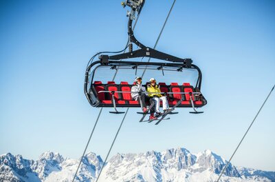 Ein Sessellift im Schnee mit zwei Personen, die hinauf in die Berge fahren. Im Hintergrund sind schneebedeckte Gipfel und ein klarer blauer Himmel zu sehen.