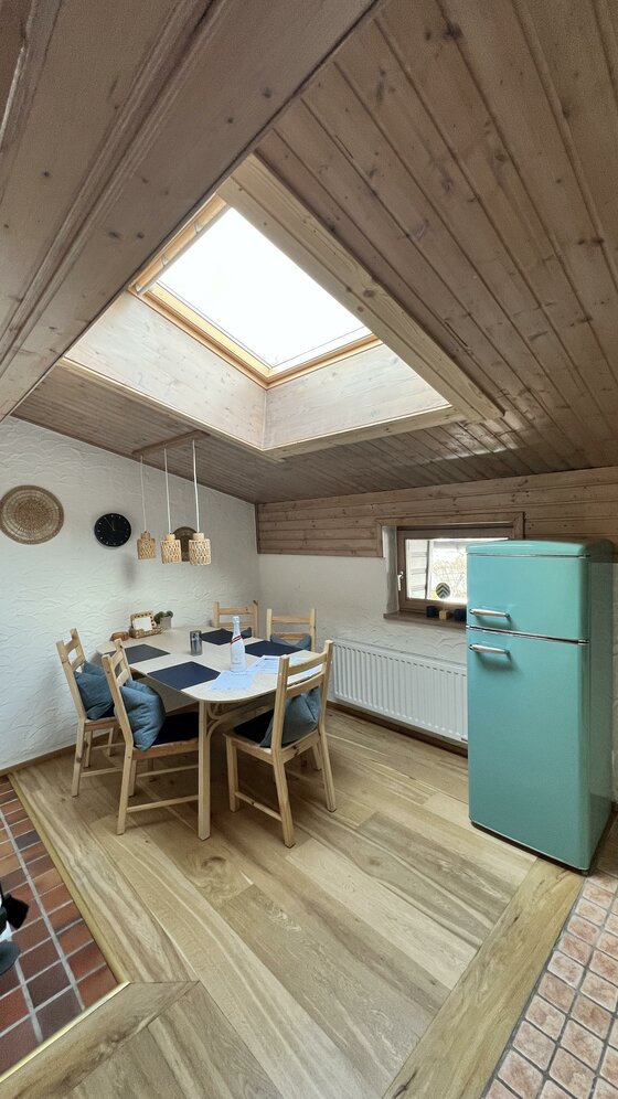 IMG_6497 Eine gemütliche Küche mit einem großen Tisch und Holzstühlen. Ein blauer Kühlschrank steht neben einem Fenster mit Dachluke.