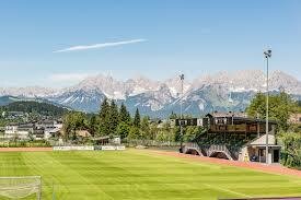 Fußballplatz mit Tribüne vor einem schneebedeckten Bergmassiv unter blauem Himmel.