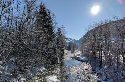 Ein flacher Flusslauf zwischen verschneiten Bäumen unter blauem Himmel mit Sonnenschein.