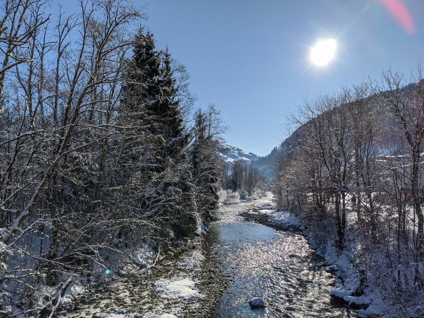 Ein flacher Flusslauf zwischen verschneiten Bäumen unter blauem Himmel mit Sonnenschein.