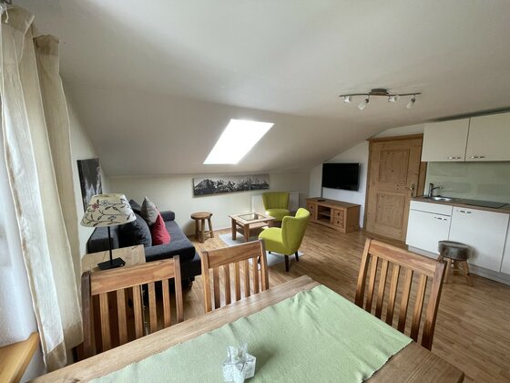 Wohnbereich mit Sofa, Sesseln, Esstisch und kleiner Küchenzeile unter einer Dachschräge mit Dachfenster.