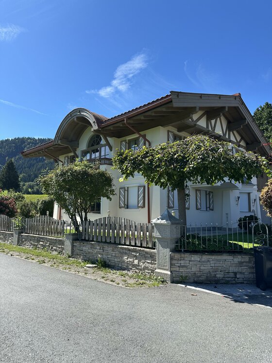 Ein schönes Haus mit einem großen Dach und einem gepflegten Garten. Im Hintergrund sind Hügel und ein blauer Himmel sichtbar.