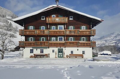 Winteransicht Angererhof Großes, mehrstöckiges Holzhaus mit Balkonen in einer verschneiten Winterlandschaft unter blauem Himmel.