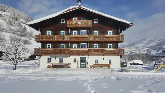 Großes, mehrstöckiges Holzhaus mit Balkonen in einer verschneiten Winterlandschaft unter blauem Himmel.