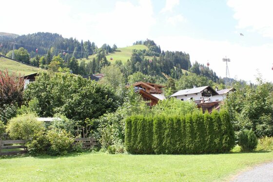 Blick über eine Wiese und Hecke auf Häuser vor einem bewaldeten Berghang mit Gondelbahn.