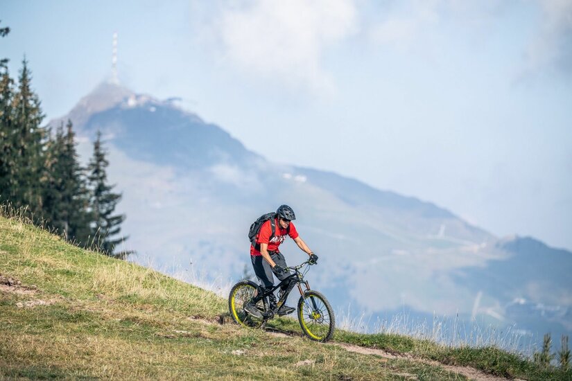 Mountainbiker am Hahnenkamm Ein Mountainbiker fährt auf einem schmalen Graspfad bergab vor einer alpinen Bergkulisse mit Sendemast.