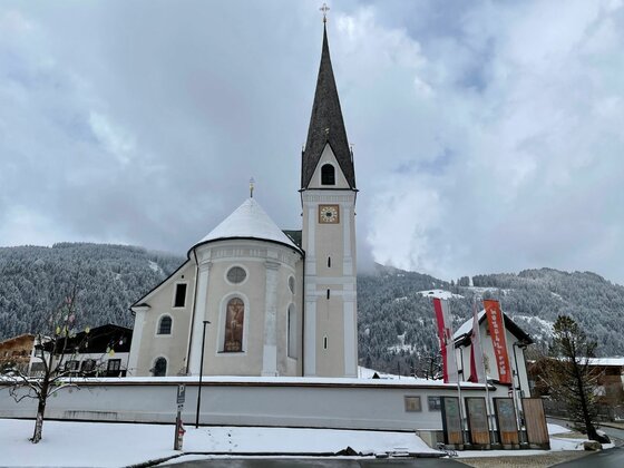 Weiße Kirche mit spitzem Turm vor verschneitem Waldhang unter bewölktem Himmel.