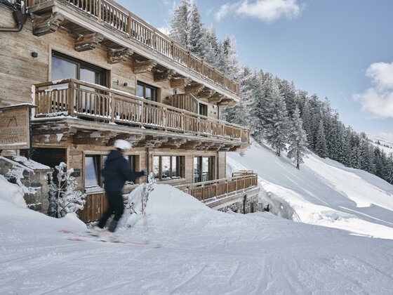 Hahnenkamm Lodge Kitzbühel Mehrstöckige Holzlodge an einer verschneiten Skipiste mit einem Skifahrer im Vordergrund.