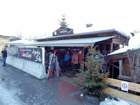 Hahnenkamm Pavillon Außenansicht Außenansicht einer Après-Ski-Bar mit abgestellten Skiern und Gästen unter einer Markise.