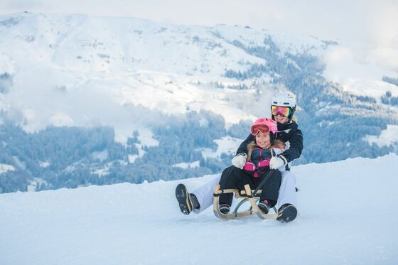 Rodeln am Gaisberg Zwei Personen fahren gemeinsam auf einem Holzschlitten eine verschneite Piste hinunter.