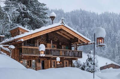 Verschneites Holzhaus in den Alpen mit Laternen und Tannenwald im Hintergrund.