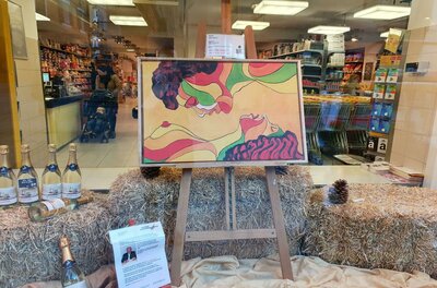 Ein buntes Kunstwerk in einem Schaufenster, das zwei Profilgesichter zeigt. Darunter stehen Flaschen und dekoratives Stroh.