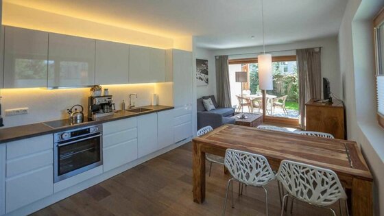 Wohnraum Kitz Apartment Moderne weiße Einbauküche mit angrenzendem Holzesstisch und Blick in den Wohnbereich.