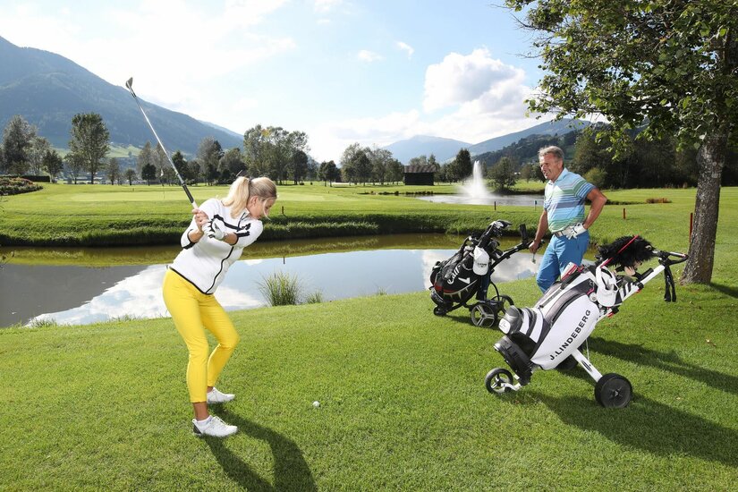 Eine Frau beim Golfabschlag auf einem grünen Golfplatz mit Wasserhindernis und Bergen im Hintergrund.