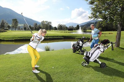 Golfen im Nationalpark Hohe Tauern Eine Frau beim Golfabschlag auf einem grünen Golfplatz mit Wasserhindernis und Bergen im Hintergrund.