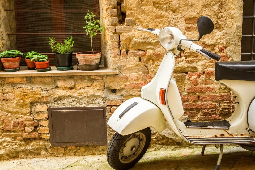 Weißer Motorroller vor einer rustikalen Ziegelwand mit Kräutertöpfen auf einem Fenstersims.