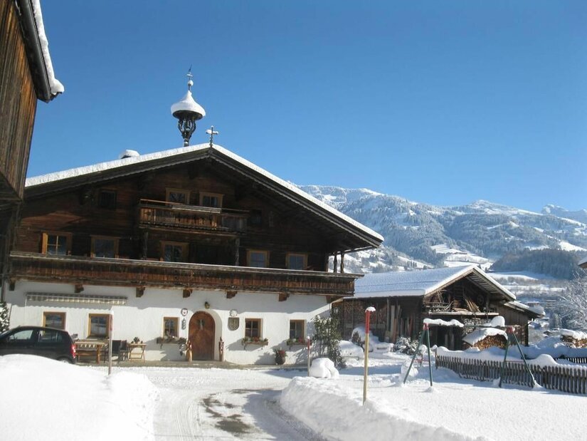 Malernhof Kitzbühel im Winter Traditionelles Bauernhaus aus Holz und weißem Putz in einer verschneiten Gebirgslandschaft unter blauem Himmel.