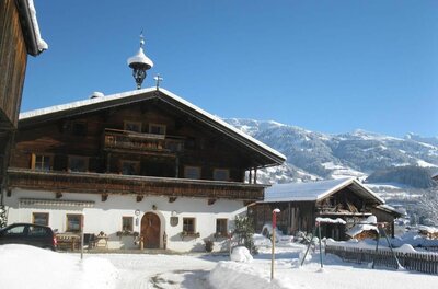 Malernhof Kitzbühel im Winter Traditionelles Bauernhaus aus Holz und weißem Putz in einer verschneiten Gebirgslandschaft unter blauem Himmel.