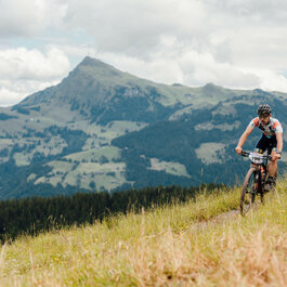 In Kitzbühel, a mountain biker rides along a narrow trail through a green meadow, with mountains in the background and clouds overhead.