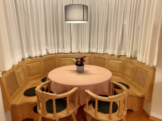 Runder Esstisch mit rosa Decke, umgeben von einer hölzernen Eckbank und zwei Stühlen vor weißen Vorhängen.