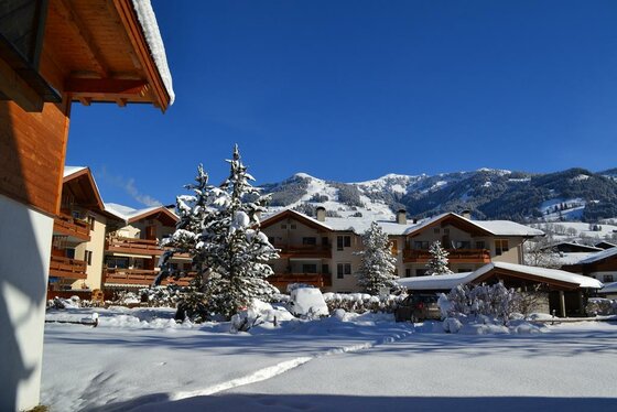 Verschneite Hotelanlage mit Holzbalkonen vor bewaldeten Bergen unter blauem Himmel.