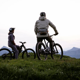 Two mountain bikers stand on a hill at sunrise, looking out over the surrounding mountains at the Hahnenkamm in Kitzbühel