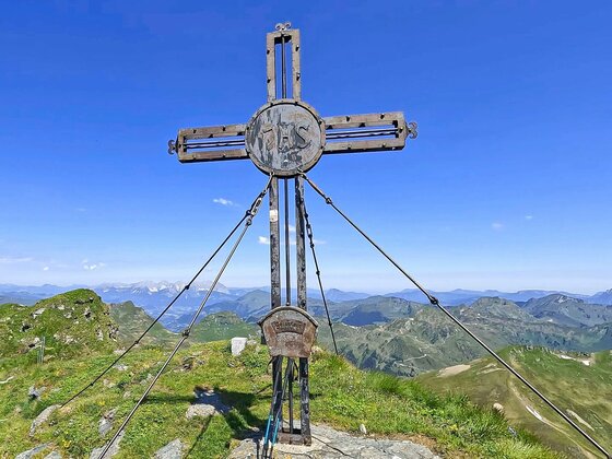 Gipfelkreuz am Geisstein Ein metallisches Gipfelkreuz auf einem grasbewachsenen Berggipfel vor blauem Himmel.