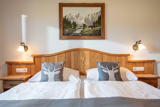Hotel_Zimmermann_Griesbachweg_48_Reith_bei_Kitzbue Ein gemütliches Hotelzimmer mit zwei Betten und Holzmöbeln. An der Wand hängt ein Landschaftsbild, und auf den Kissen sind Hirschmotive.