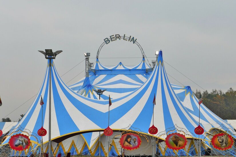 Blau-weiß gestreiftes Zirkuszelt mit dem Schriftzug Berlin auf einem Metallbogen vor grauem Himmel.
