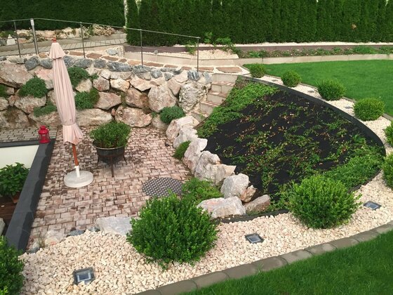 Gartenanlage Appartement Lyso Gepflasterte Terrasse mit Sonnenschirm, Natursteinmauer und bepflanzten Hangbeeten.