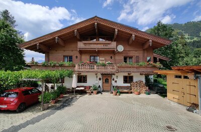 Alpines Holzhaus mit Blumenbalkon, gepflastertem Vorplatz und einem roten Auto unter einer Pergola.