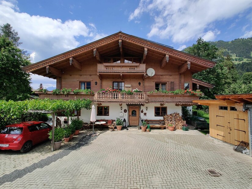Haus Berger Alpines Holzhaus mit Blumenbalkon, gepflastertem Vorplatz und einem roten Auto unter einer Pergola.