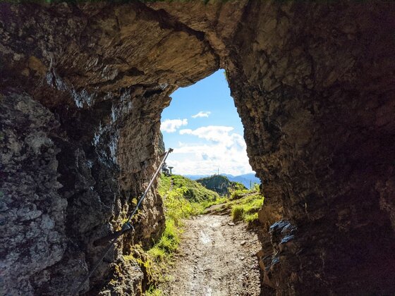 Felstunnel am Horn Gipfel-Höhenweg Blick aus einem dunklen Felstunnel auf einen sonnigen Bergpfad mit Drahtseilsicherung unter blauem Himmel.