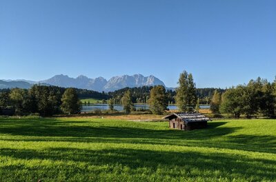 Schwarzsee mit Kaisergebirge Grüne Wiese mit kleiner Holzhütte vor einem See und Gebirgskette unter blauem Himmel.