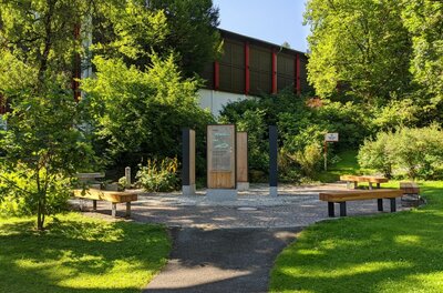Infoplatz Streifzug durch Kitzbühel Gepflasterter Platz mit hölzernen Infostelen und Sitzbänken inmitten von Bäumen und Sträuchern.