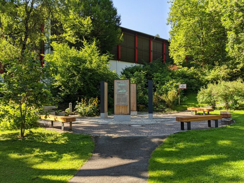 Infoplatz Streifzug durch Kitzbühel Gepflasterter Platz mit hölzernen Infostelen und Sitzbänken inmitten von Bäumen und Sträuchern.