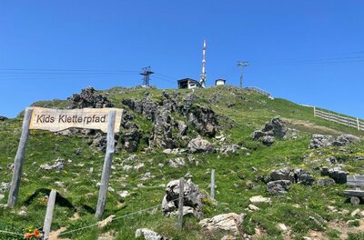 Holzschild mit der Aufschrift Kids Kletterpfad auf einem grasbewachsenen Berghang unter blauem Himmel.
