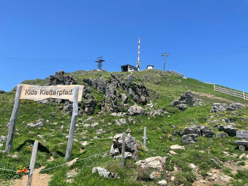 Kids Kletterpfad am Kitzbüheler Horn Holzschild mit der Aufschrift Kids Kletterpfad auf einem grasbewachsenen Berghang unter blauem Himmel.
