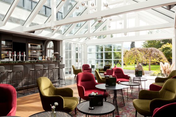 Greenhouse Bar im Hotel Erika Moderner Wintergarten mit Bar, roten und grünen Sesseln sowie Blick in den Garten.