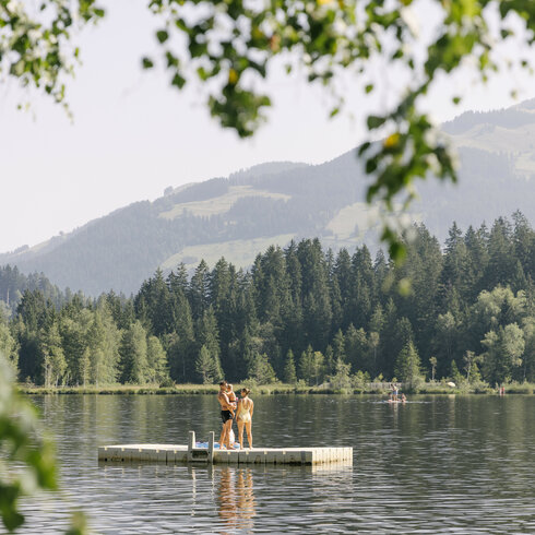 Eine Familie genießt einen sonnigen Tag auf einer schwimmenden Insel am Schwarzsee in Kitzbühel und bewundert die Natur mit dem Kitzbüheler Horn im Hintergrund.