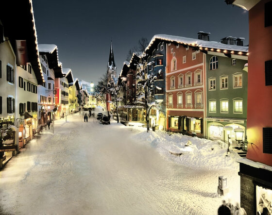 Kitzbühel im Winter Verschneite Fußgängerzone in Kitzbühel bei Nacht mit beleuchteten bunten Häuserfassaden und Kirchturm.