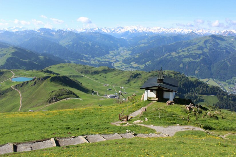 Kitzbüheler Horn Eine malerische Berglandschaft mit grünen Wiesen und schneebedeckten Gipfeln im Hintergrund. Im Vordergrund steht eine kleine Kapelle auf einem Hügel.