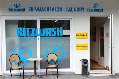 Ein Waschsalon mit der Aufschrift "Kitzwash". Vor dem Eingang stehen zwei Stühle und ein kleiner Tisch.